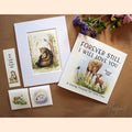 A styled flat-lay of a children's memory book and matching keepsake items on a brown surface. The book cover reads 'FOREVER STILL, I WILL LOVE YOU' and shows an illustration of a bear and deer.